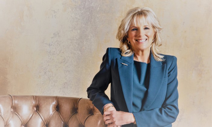 Dr. Jill Biden wears a blue suit, standing next to leather armchair